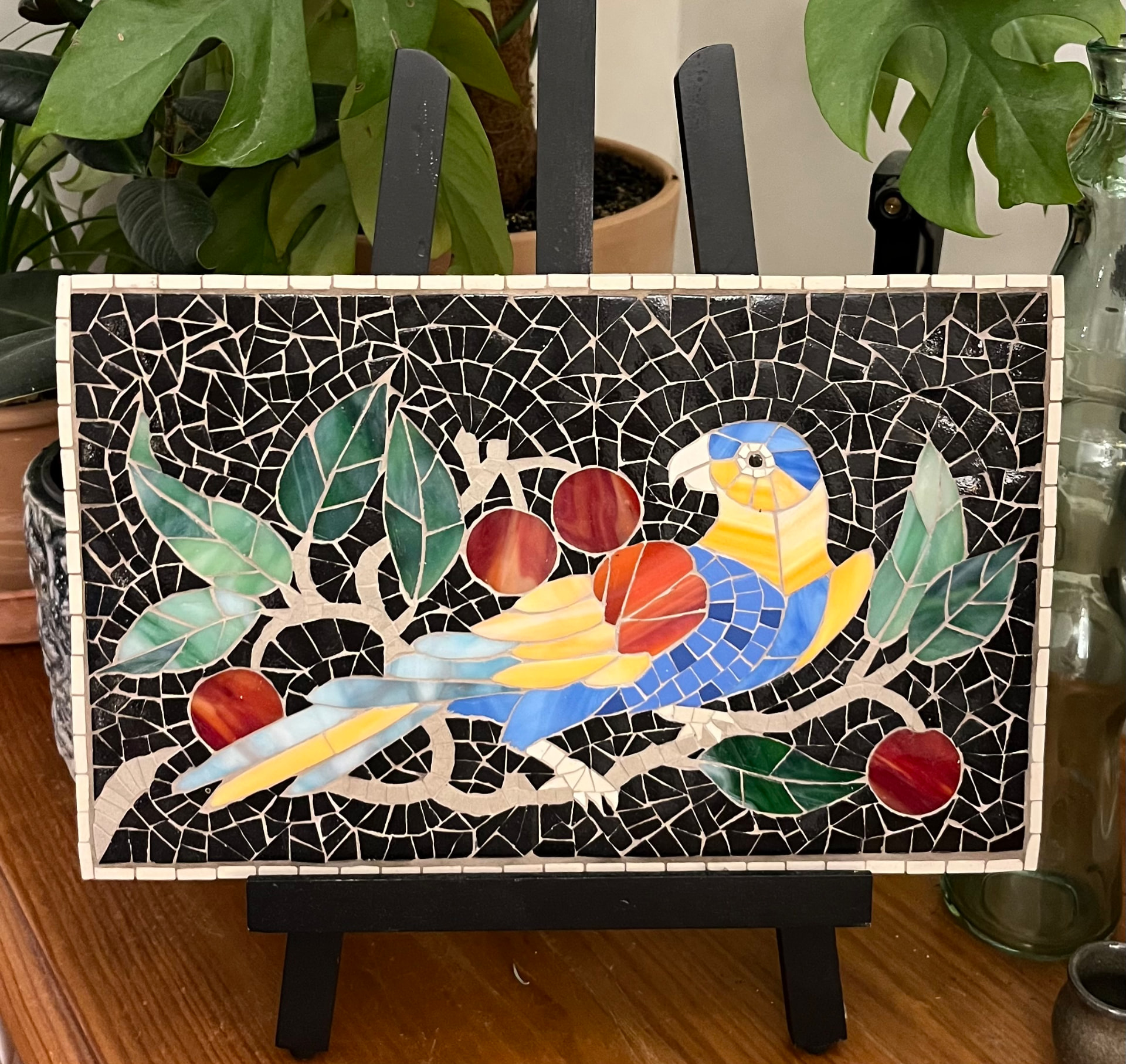 Tableau en mosaïque représentant un perroquet bleu et jaune entouré de feuillages sur fond noir