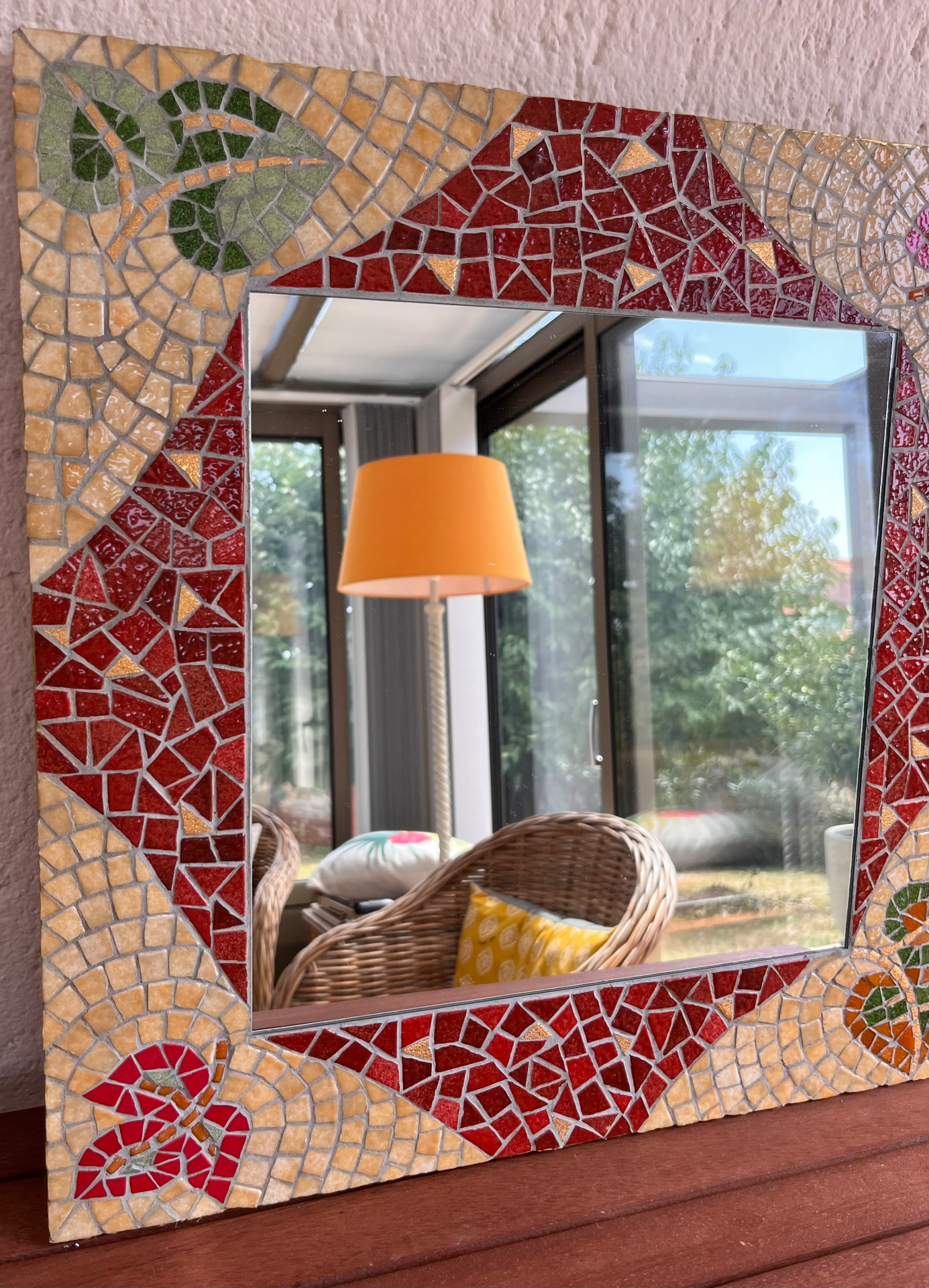 Miroir en mosaïque rouge, or et vert à motifs de feuilles et de libellule, reflétant un intérieur lumineux