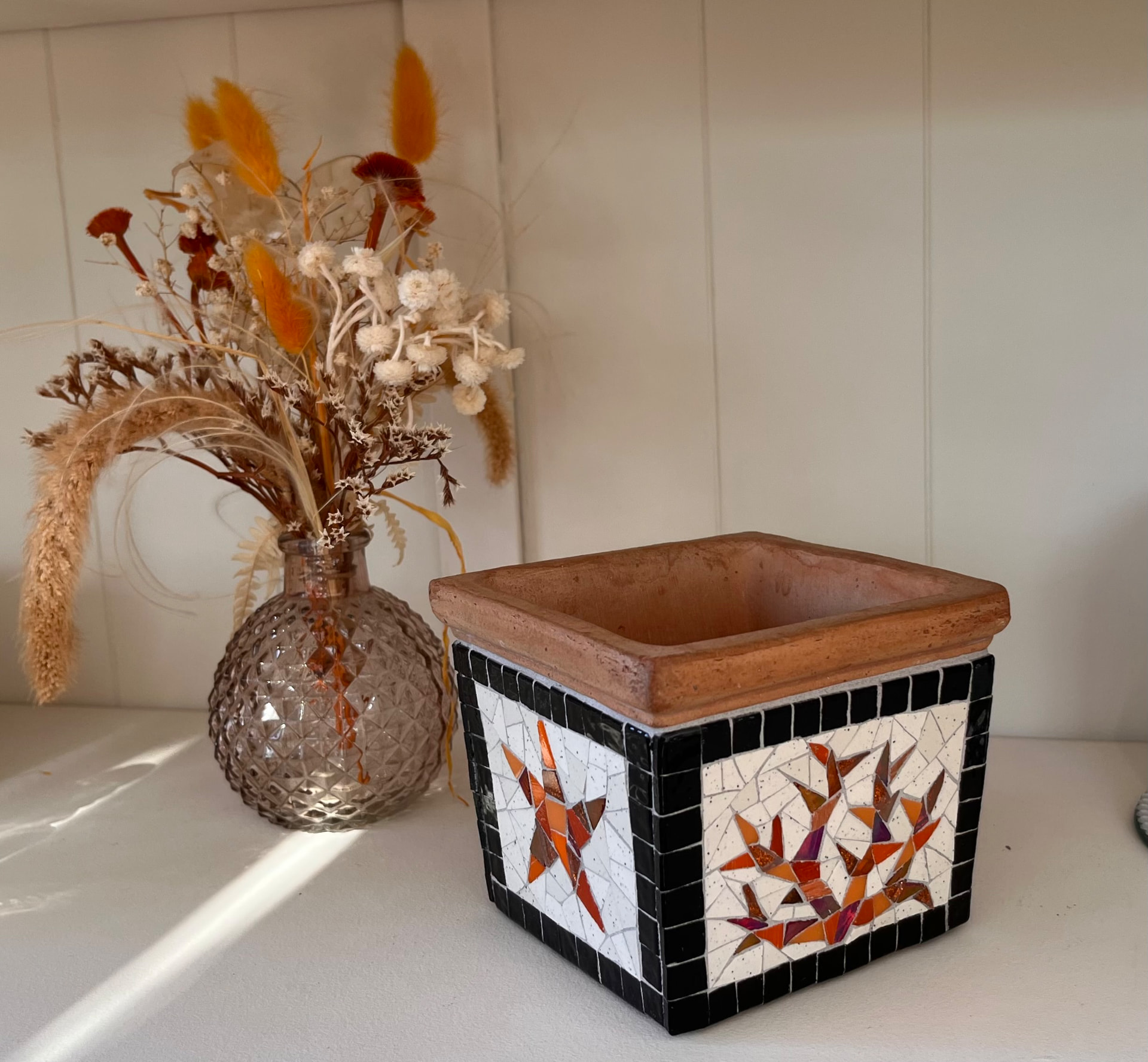 Pot carré en terre cuite décoré d’une mosaïque à motif d’oiseaux orange et noir, à côté d’un vase en verre et de fleurs séchées
